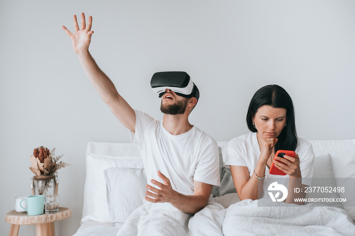 Serious young adult woman sitting on bed holds phone watching news while her boyfriend playing games using virtual reality headset. Couple in bed at morning. Bored woman using smartphone. Disunity