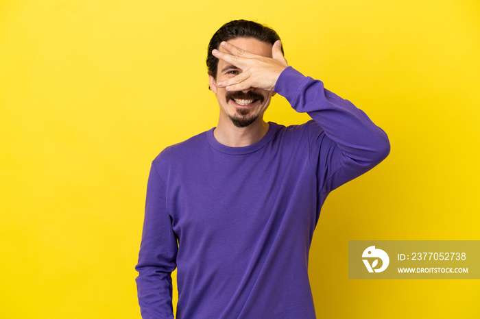 Young caucasian man isolated on yellow background covering eyes by hands and smiling