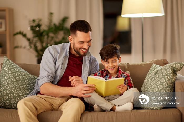 family, childhood, fatherhood, leisure and people concept - happy smiling father and little son reading book on sofa at home in evening