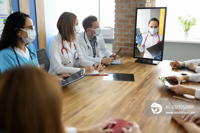 Medical staff sit at table with documents and look at monitor on which doctor sees conversation. Online medical meetings and consultations concept