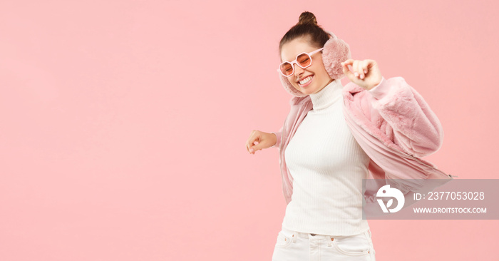 Horizontal banner of young active girl wearing fluffy winter coat, ear warmers and colored glasses, dancing to music, isolated on pink background with copy space