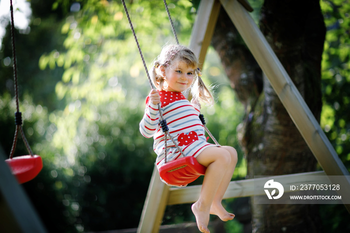 Happy beautiful little toddler girl having fun on swing in domestic garden. Cute healthy child swinging under blooming trees on sunny spring day. Baby laughing and crying