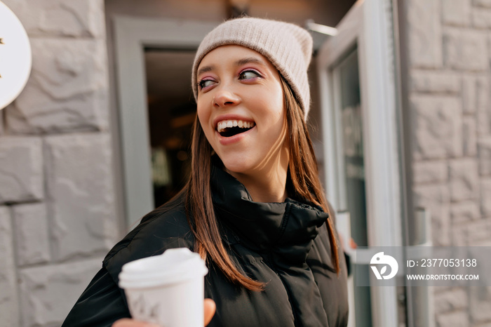 Excited lovely girl with great smile in knitted cap coming out from cafe with coffee to go. Pretty charming lady walking around the city in sunny warm spring day