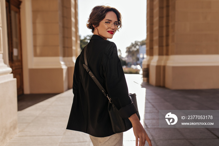 Wonderful girl in black jacket with dark handbag smiling at street. Short-haired woman in eyeglasses with red lips posing outside..