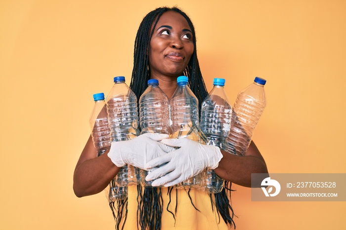 African american woman with braids holding recycling plastic bottles smiling looking to the side and staring away thinking.