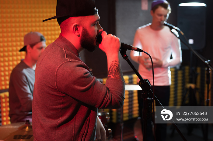 Side view portrait of tattooed hip-hop musician performing in dim recording studio while making new album with his band, copy space