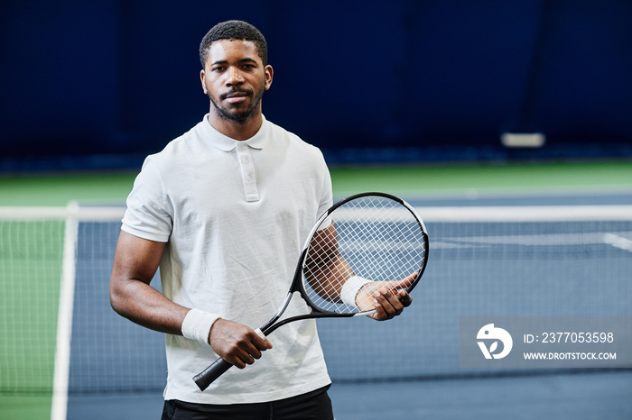 Waist up portrait of African American tennis player looking at camera while posing confidently with racket at indoor court, copy space