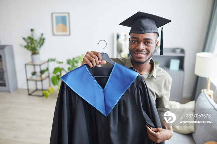 Portrait of happy African-American man holding graduation gown and smiling at camera while preparing for ceremony at home, copy space