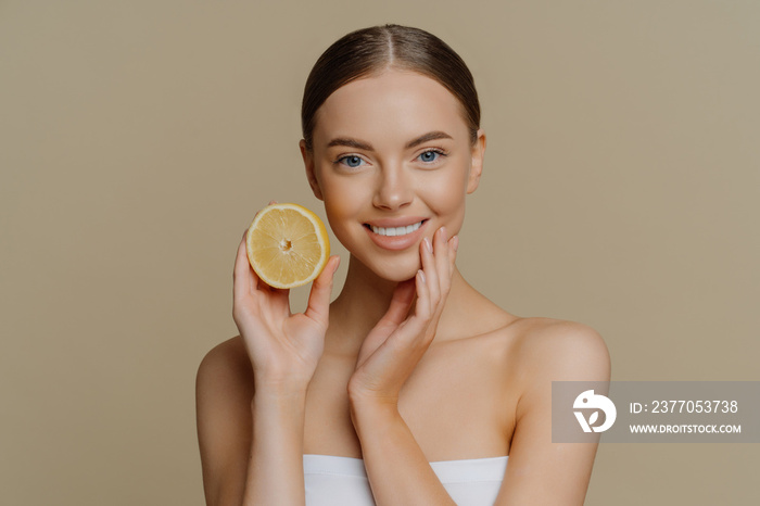 Healthy glad young woman poses with bare shoulders indoor wrapped in bath towel holds slice of lemon smiles tenderly has minimal natural makeup isolated over brown background. Skin care concept