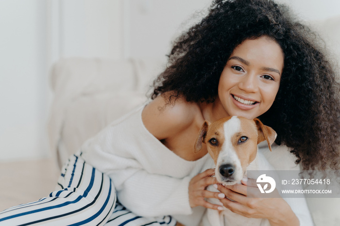 Image of attractive dark skinned woman wears makeup, has bushy curly hair, smiles pleasantly, cuddles dog, dressed in fashionable clothes, enjoys sweet moment with puppy, being at home together