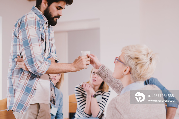 Young addicted people having stressed situation while sitting together on special group therapy. Upset bearded guy taking water to calm down after his confession.