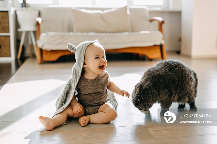 Happy baby sitting on the floor playing with a cat, wearing a hooded cardigan with ears, laughing