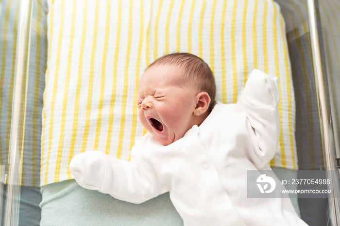 Newborn baby yawning, lying in a hospital crib, first days after birth, motherhood concept.