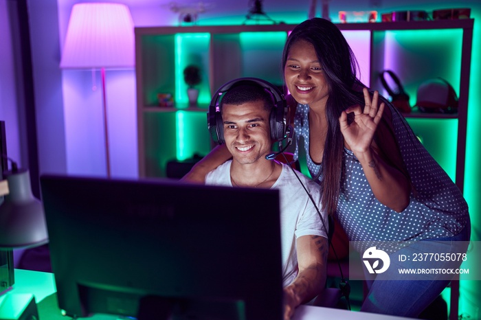 Young hispanic couple playing video games doing ok sign with fingers, smiling friendly gesturing excellent symbol