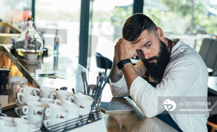 Worried Barista Using Digital Tablet in a Cafe.  Distraught sad waiter with a beard leaning on the bar counter and looking away while waiting for order in the restaurant or a coffee shop.