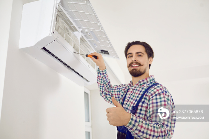 Portrait of handsome mustached AC repair service worker holding screwdriver, looking at camera with happy face expression and showing thumbs up while fixing air conditioner unit at home or in office