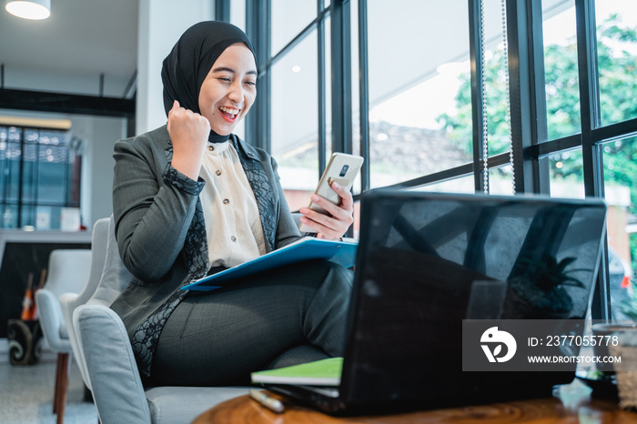 portrait of happy excited muslim woman raise her arm while looking at her phone