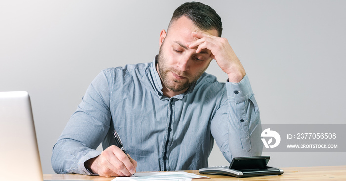 Unhappy accountant businessman stressfuly filling tax form with calculator at office desk.