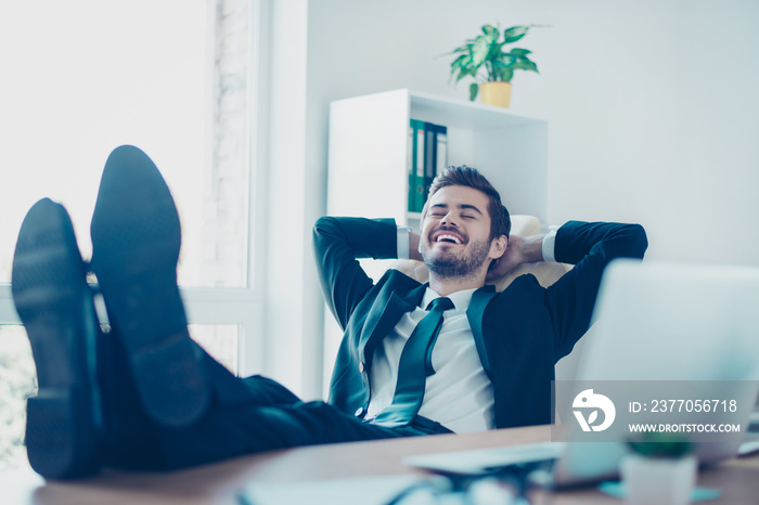 Happy cheerful smiling young entrepreneur  lying on the chair with legs on desktop at office and having a rest after solving all tasks