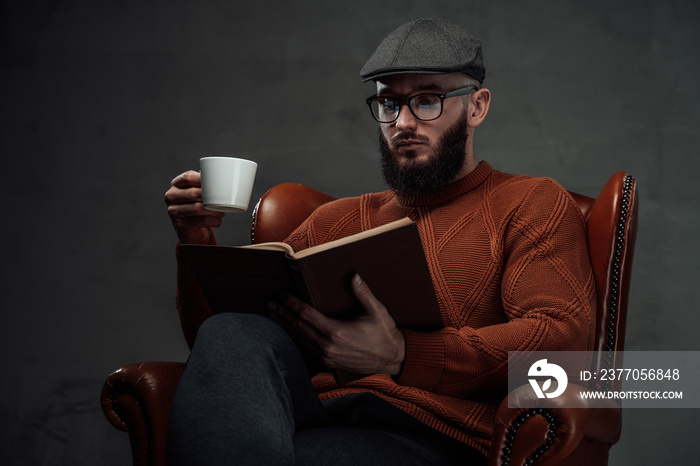 Sitting on leather armchair in dark background clever and manly guy with beard and glasses weared in sweater with cap.