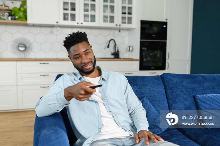 Happy young multiracial man holding tv remote control while sitting on a couch at home and relaxing at the weekend. Stock photo