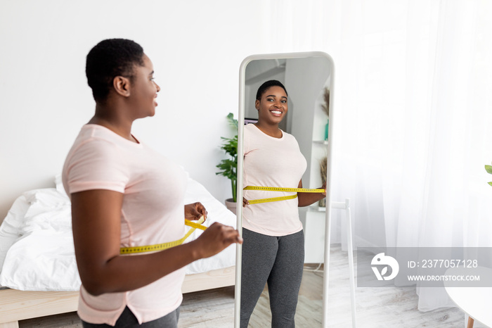 Overweight young black woman measuring waist with tape near mirror at home, satisfied with slimming result, free space