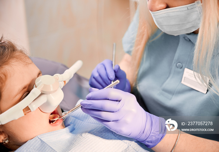 Female dentist checking child teeth with dental explorer and mirror while girl lying in dental chair with inhalation sedation at dental office. Concept of pediatric, sedation dentistry.