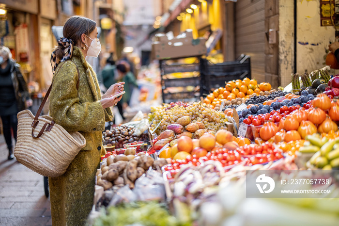 Woman in medical mask buying fruits and vegetables at market stall on the famous gastronomical street in Bologna. Concept of buying local products during pandemic. Idea of Italian lifestyle