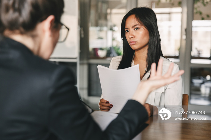 Image of serious asian woman looking and talking to businesswoman, while sitting at table in office during job interview - business, career and recruitment concept