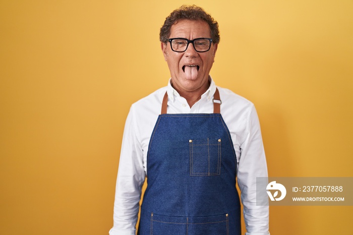 Middle age hispanic man wearing professional cook apron sticking tongue out happy with funny expression. emotion concept.