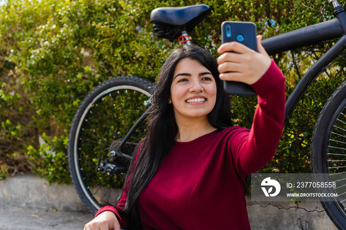 Young hispanic woman sitting on a stair with her bike behind her and a bush taking a selfie