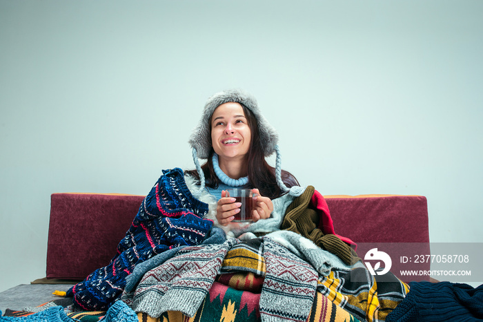 The young sick woman with flue sitting on sofa at home or studio covered with knitted warm clothes. Illness, influenza, pain concept. Relaxation at Home. Healthcare Concepts.