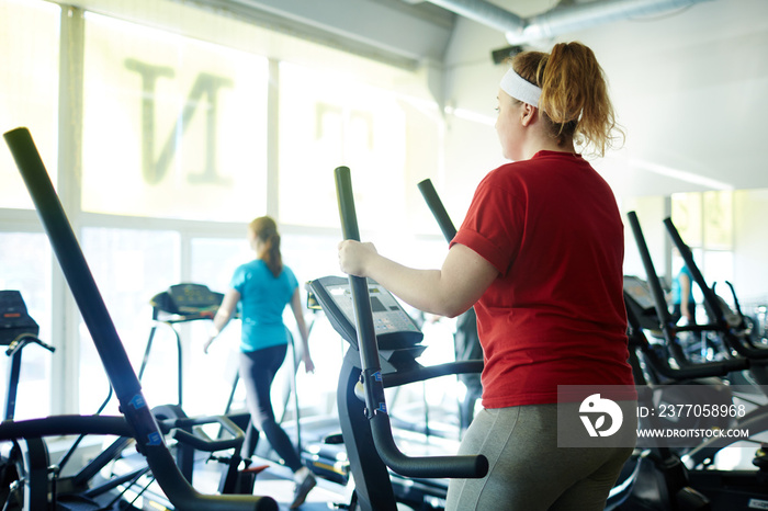 Back view  portrait of young obese woman working out in gym: using ellipse  machine with effort to lose weight