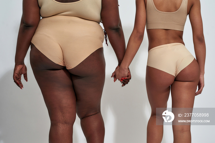 Cropped shot of two african american women in underwear with different body size holding each other hands, standing isolated over gray background