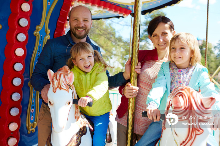 Pretty little girl in green windbreaker and her elder sister riding on carousel while their cheerful parents standing behind them and posing for photography