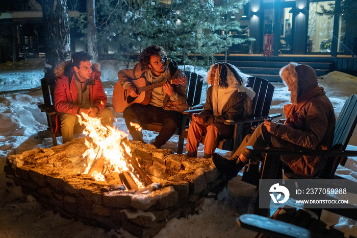 Group of friends playing guitar and singing songs together while sitting near the fire outdoors in winter evening