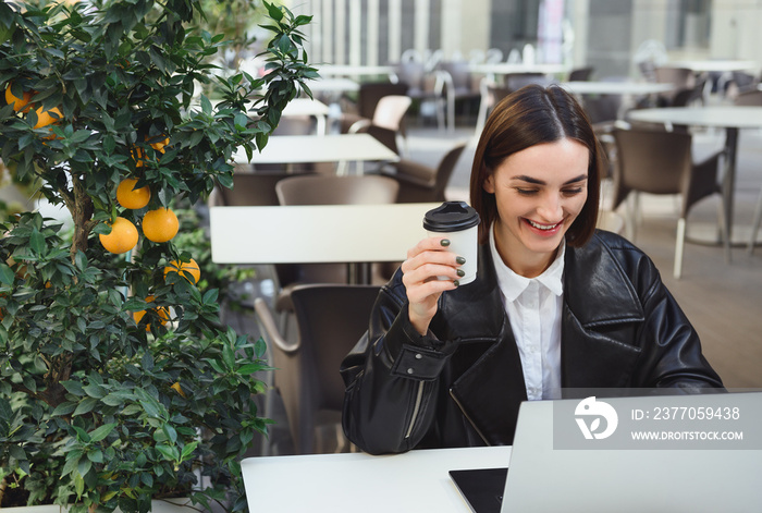 Beautiful young business woman, freelancer, developer, journalist, copywriter holds takeaway cup of coffee, works online in outdoor cafe, smiles reading good news from business partners on laptop