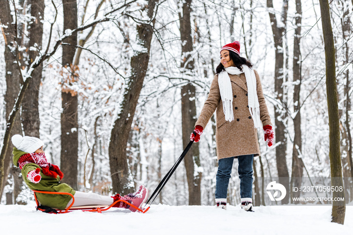 attractive african american woman pulling preteen daughter on sledge and smiling in winter park