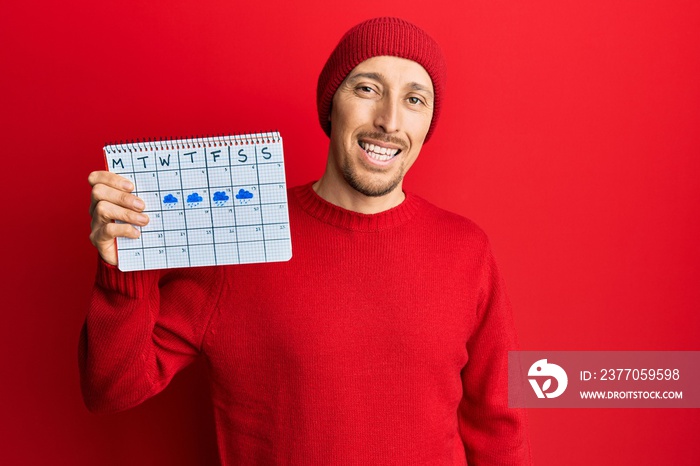 Bald man with beard holding rainy weather calendar looking positive and happy standing and smiling with a confident smile showing teeth