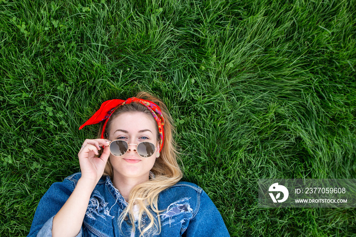 Portrait of an attractive stylish girl in a denim jacket lying on a green lawn, looking into the camera and holding hands with the sunglasses, a view from above. Copyspace