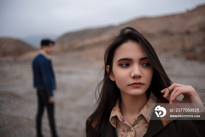 Dramatic portrait of a young brunette girl in cloudy weather. somewhere behind her, out of focus, her young lover boyfriend leaves her after break up . selective focus, small focus area