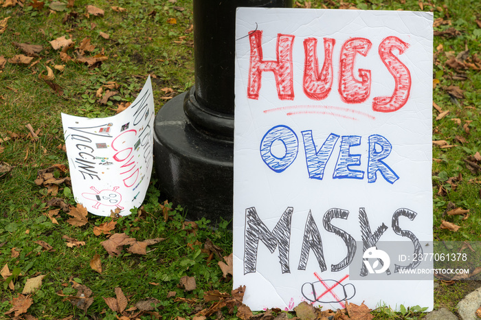 A hugs over masks sign. Drawn mask with X through it at the bottom. Another sign protesting covid vaccines to the left, but only partly readable.