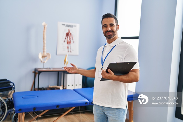 Young hispanic man physiotherapist smiling confident holding clipboard at rehab clinic