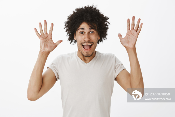 Waist-up shot of joyful carefree attractive male with curly hair in grey t-shirt, raising palms high in jazz hands gesture, smiling broadly, being in playful mood while having fun over gray wall