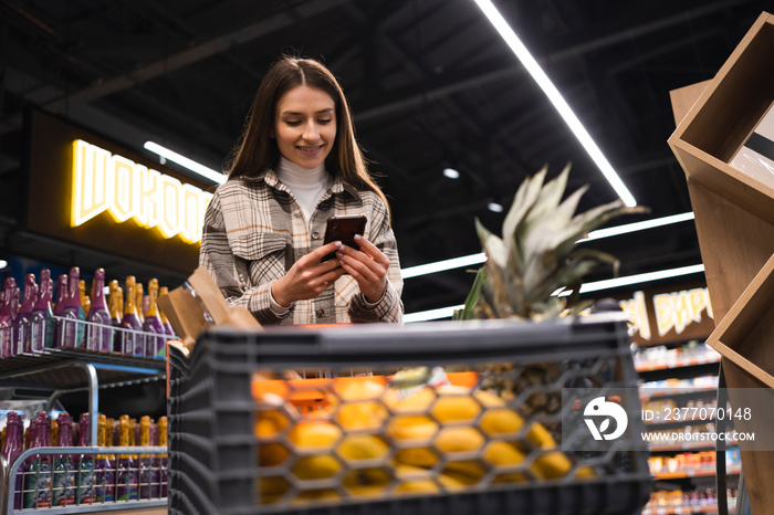 Beautiful young woman with shopping trolley looking at shopping list in smartphone.