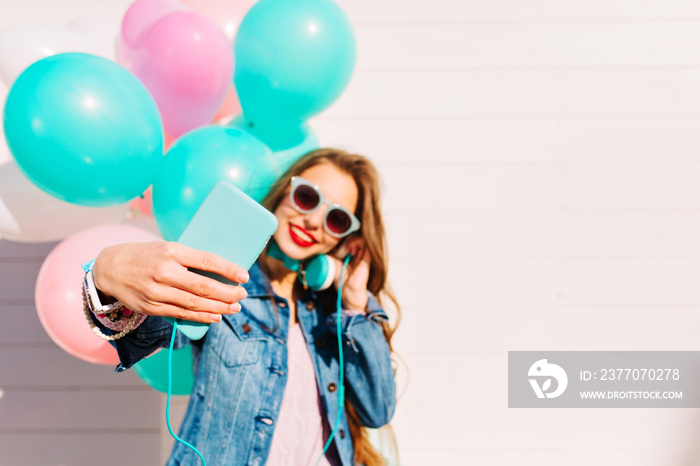 Adorable young long-haired woman with balloons makes selfie on smartphone while listening music. Portrait of happy brunette girl enjoing favourite song in headphones and taking picture of herself