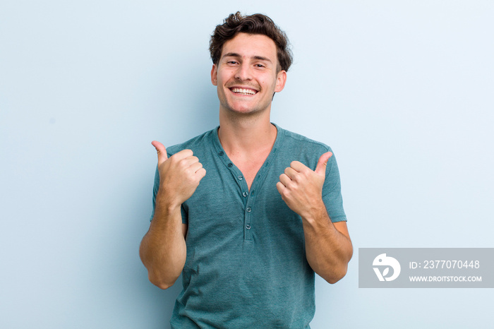 young handsome man smiling joyfully and looking happy, feeling carefree and positive with both thumbs up