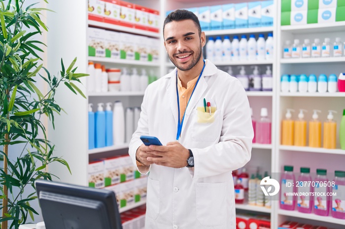 Young hispanic man pharmacist using smartphone working at pharmacy