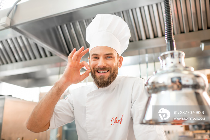 Portrait of a handsome chef cook in uniform showing delicious sign at the restaurant kitchen