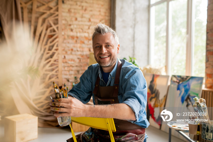 Cheerful artist in workwear sitting on chair in front of camera